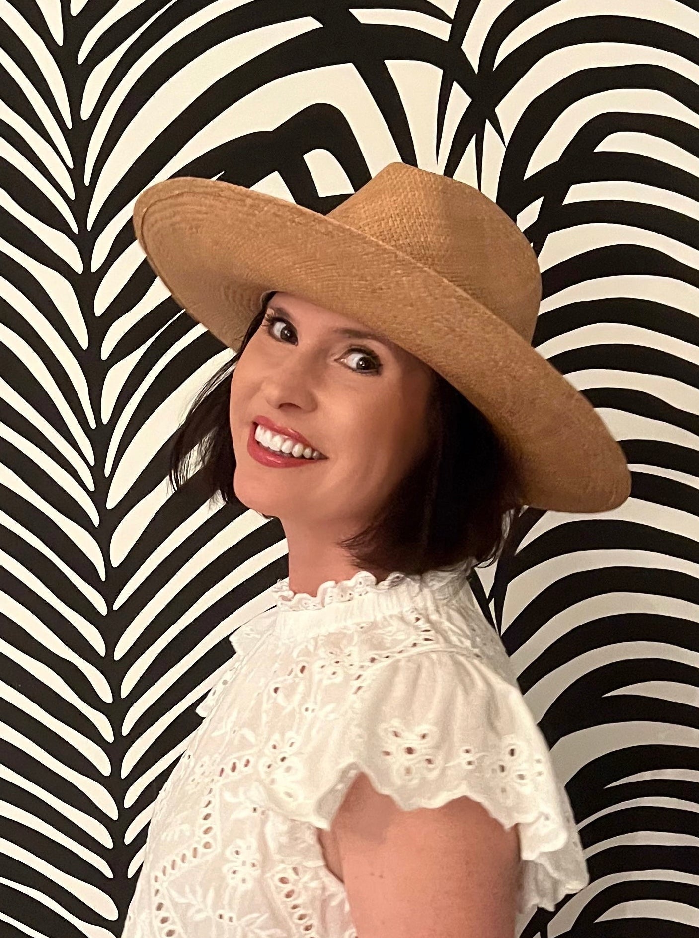 Woman in a white dress and wide-brimmed hat standing against a black and white patterned wall.