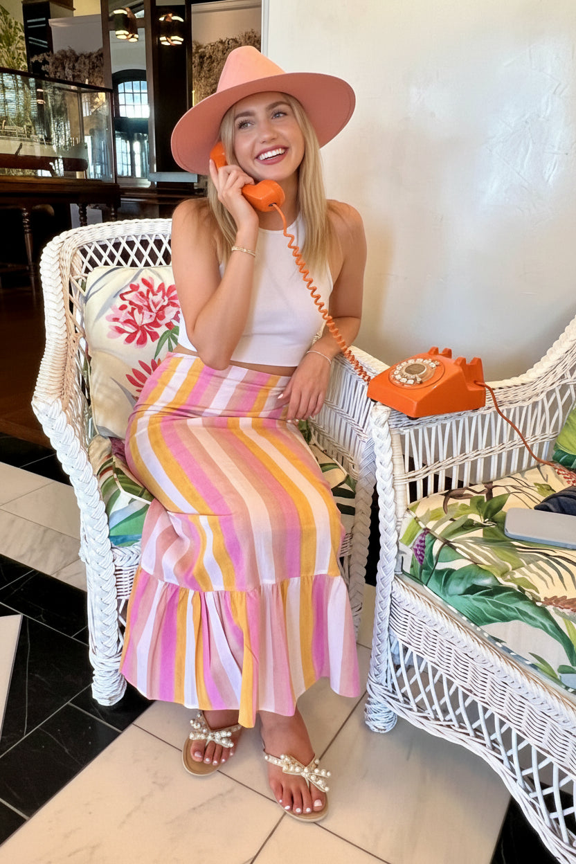 Woman sitting on a white wicker chair wearing a colorful striped skirt and pink hat.