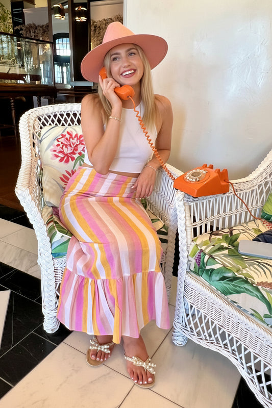 Woman sitting on a white wicker chair wearing a colorful striped skirt and pink hat.