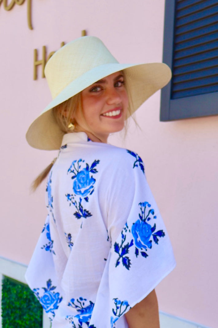 Woman wearing a white kimono with blue floral patterns and a wide-brimmed hat against a pink wall.